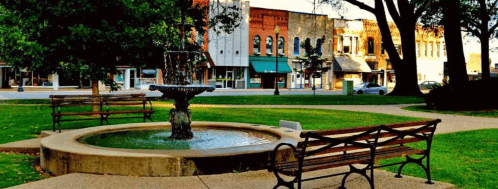 A park with a fountain surrounded by benches, with historic buildings in the background.